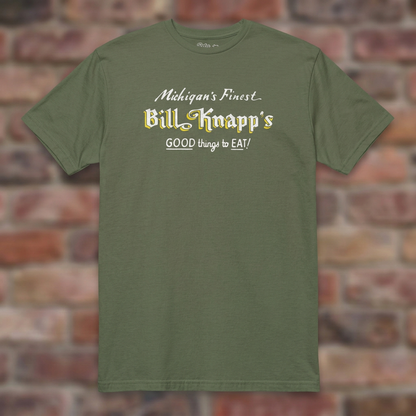 Military Green Bill Knapp’s T-shirt flat lay on warm brick background